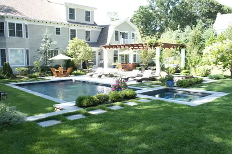 Pool and raised spa under a pergola in front of a large stone house