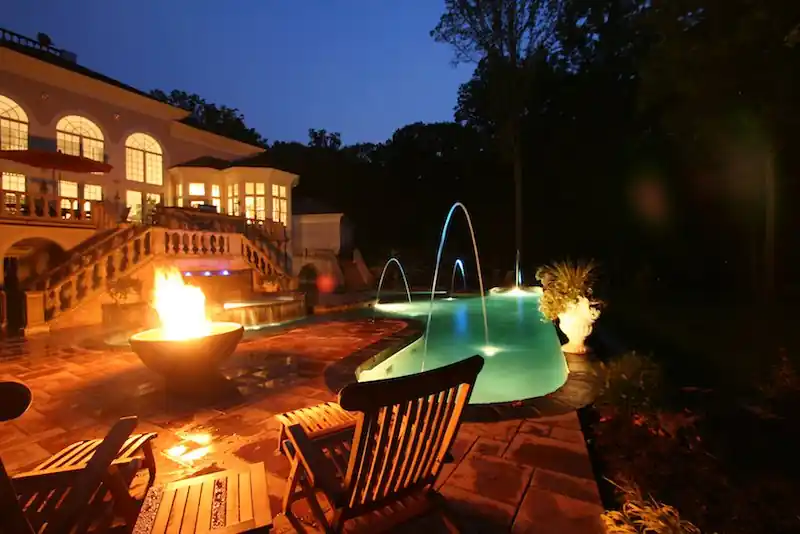 Freeform pool lit at dusk with a fire bowl and laminar jets arcing into the water
